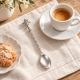 Petite cuillère Anges présentée sur du lin clair auprès d’une tasse de café et de biscuits.